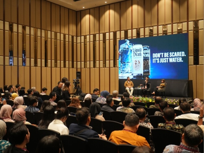 Speaker presenting to a packed audience at a professional conference, with a screen displaying the message 
