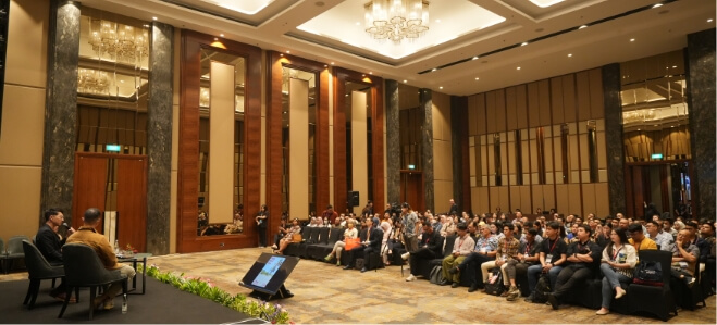 Wide-angle view of a formal conference setup with seated participants, chandeliers, and stage presentation in progress.