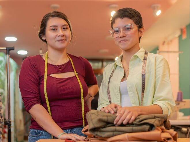 Two female fashion designers smiling at the camera in a studio, one holding fabric and both wearing measuring tapes around their necks.