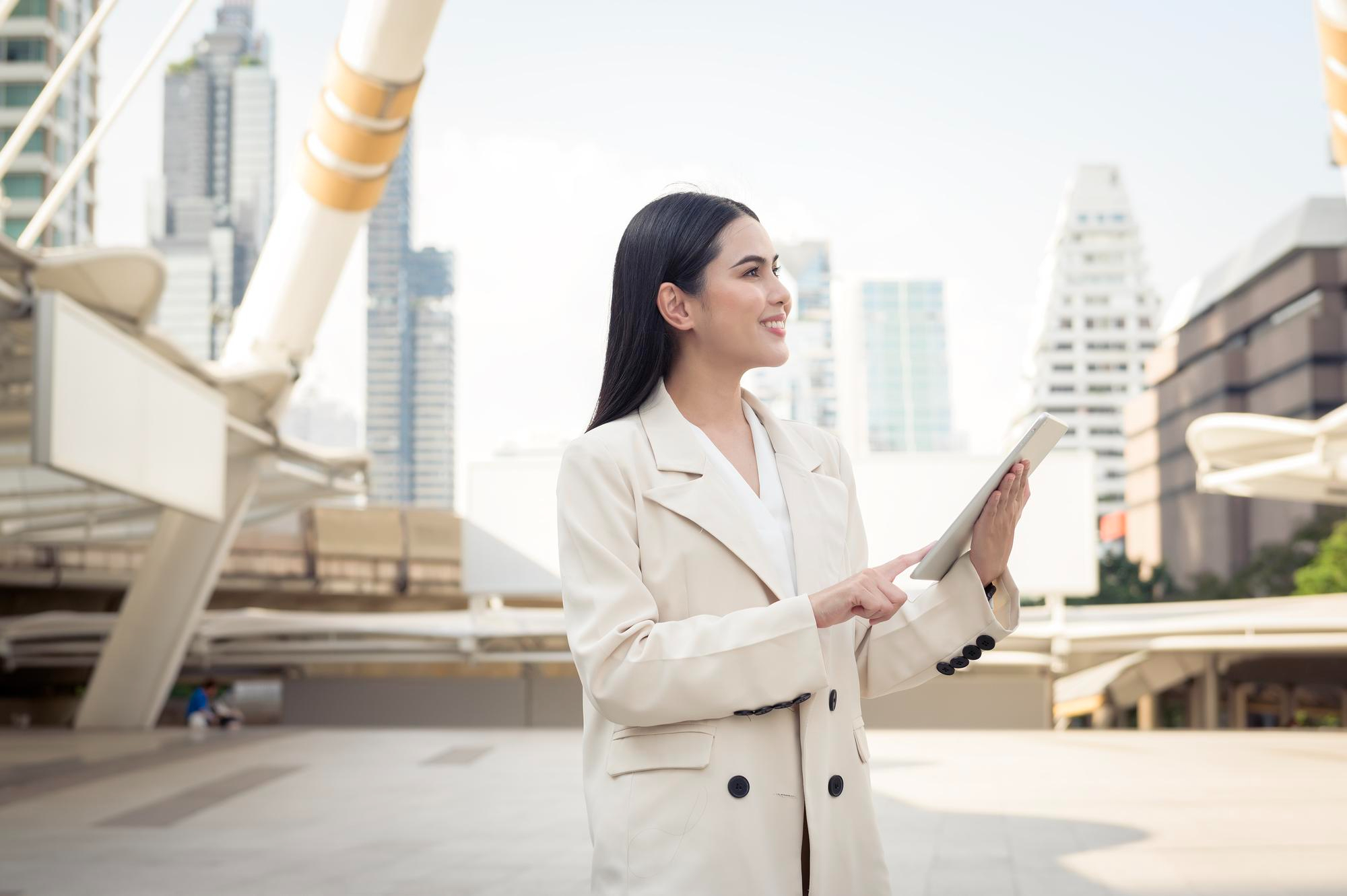 Professional Asian businesswoman using a digital tablet outdoors in a modern cityscape with high-rise buildings and a pedestrian bridge in the background.