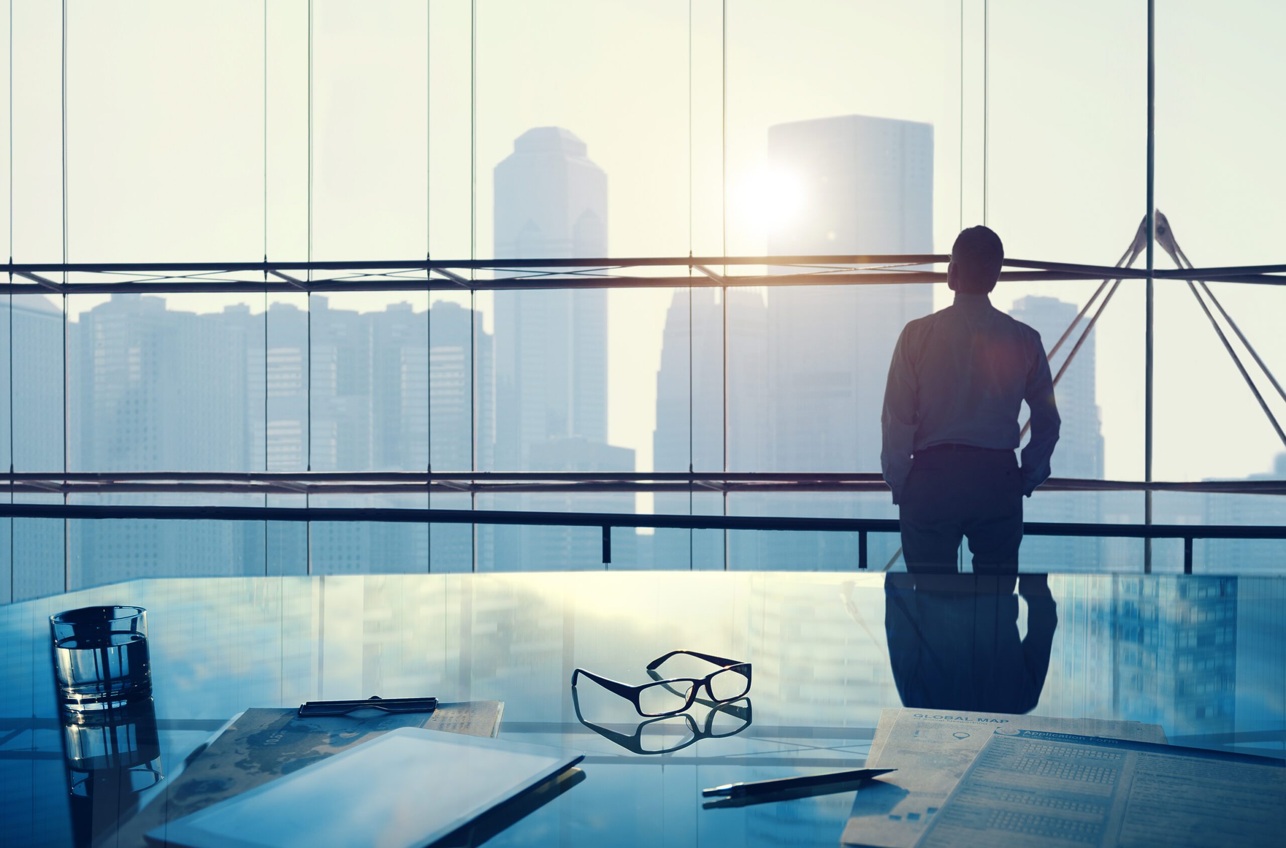 Businessman standing by a large office window overlooking a city skyline at sunrise.