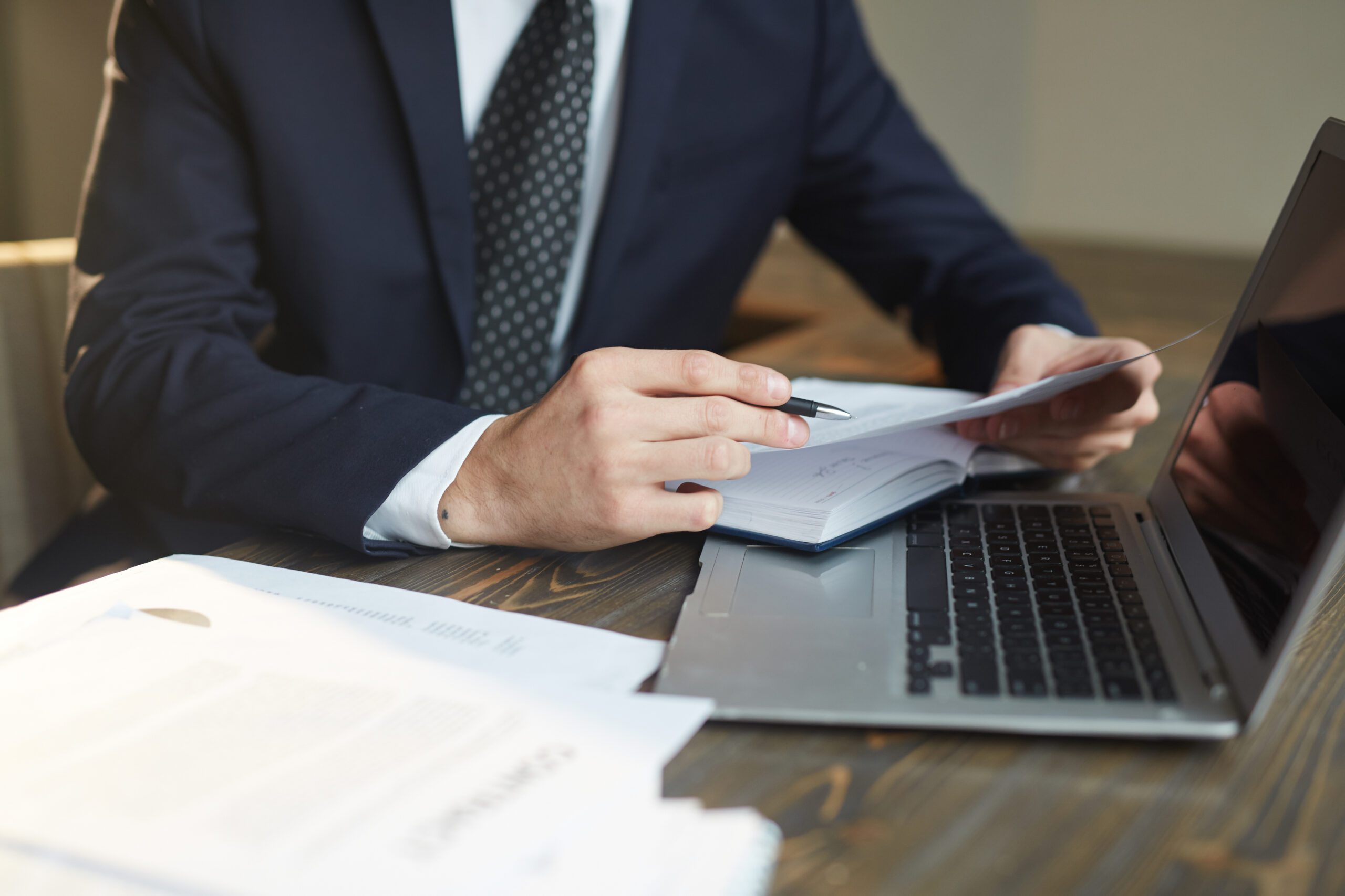 Businessman in a suit reviewing documents and taking notes while working on a laptop at a desk with papers spread out.