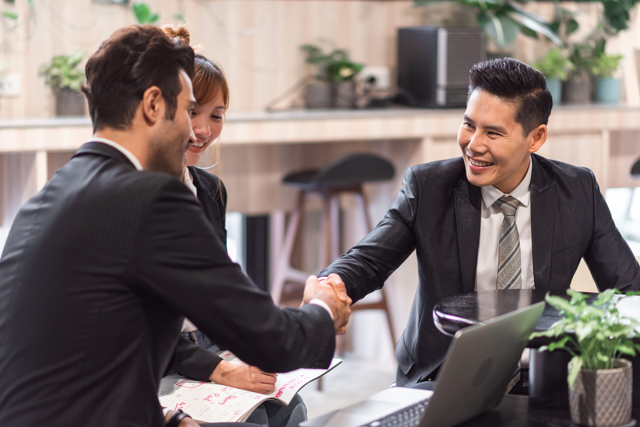 Group of business professionals in formal attire smiling and shaking hands during a meeting, symbolizing successful partnership and teamwork, with a laptop and documents on the table.