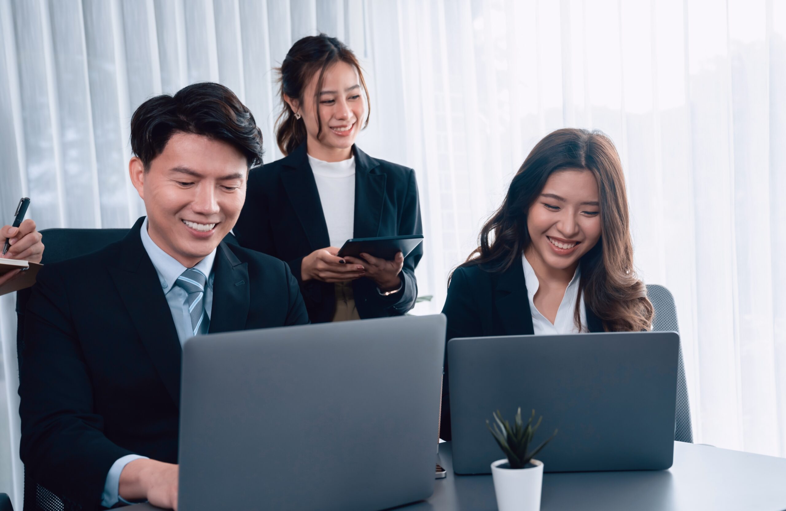 Three business professionals in formal attire smiling while working together in an office. Two are seated with laptops, while one stands holding a tablet.